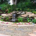 A pond with water flowing over it and flowers around.
